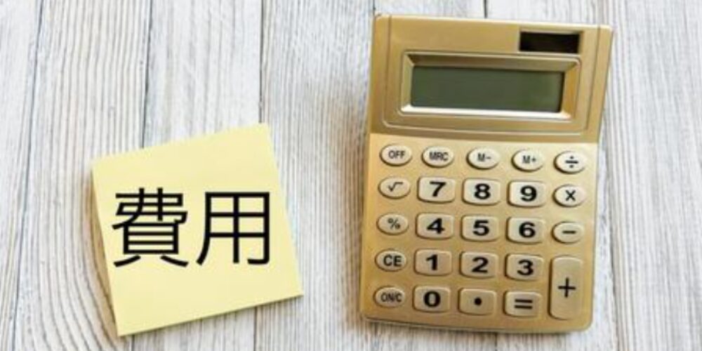 木目調の机の上に置かれた黄色の付箋に「費用」と書かれた文字と、隣にある金色の電卓