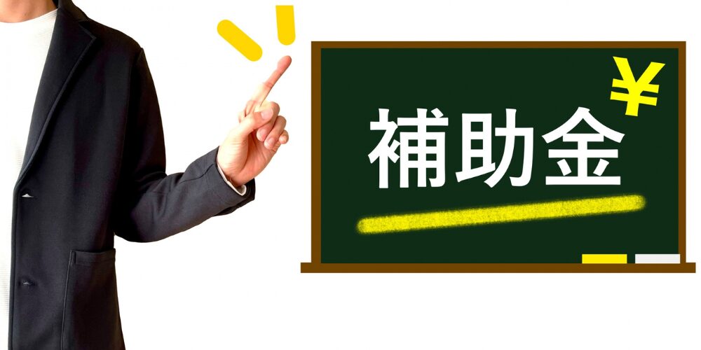 ビジネスジャケット姿の男性が『補助金』と書かれた黒板を指差している画像