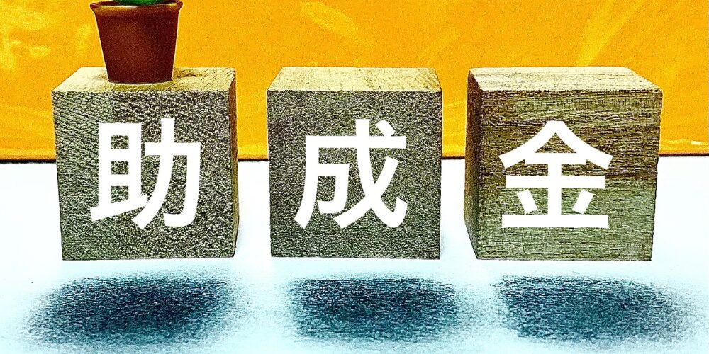 黄色い背景に木製ブロックを並べ『助成金』と示し、左端にミニ観葉植物が置かれたイメージ