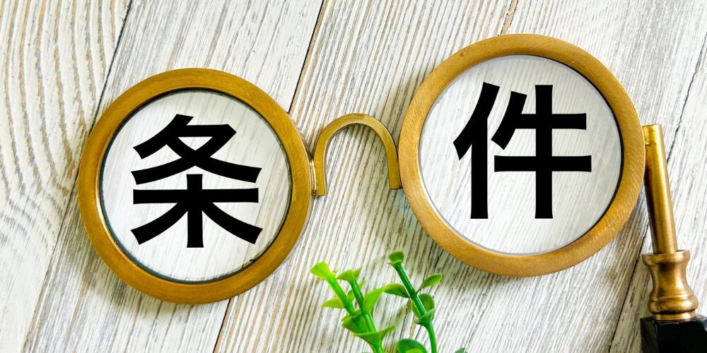 木目テーブル上で丸いルーペを眼鏡のように配置し『条件』の文字を拡大した写真
