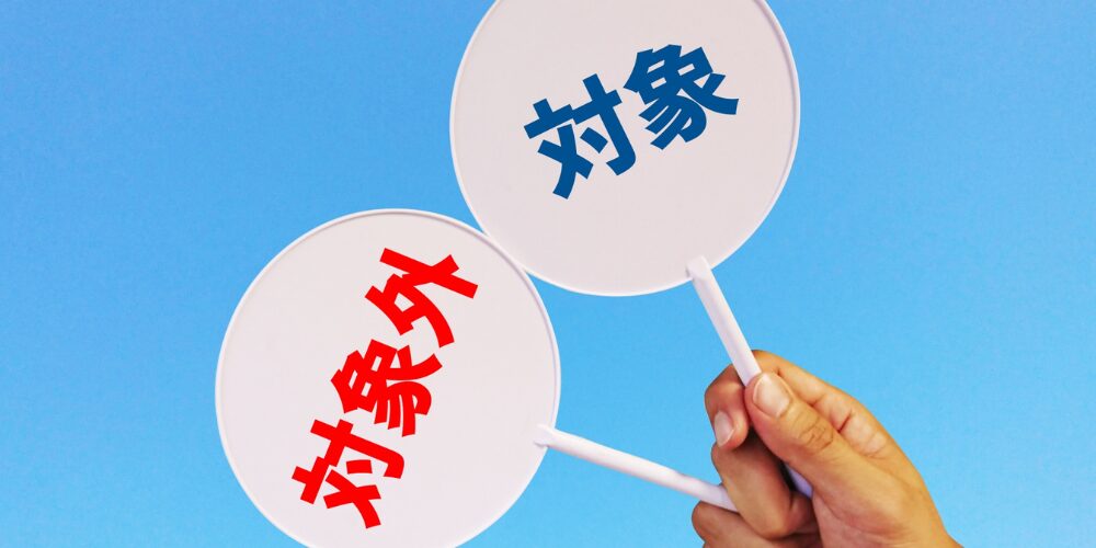 青空を背景に『対象』と『対象外』と書かれたうちわを手に持つイメージ写真