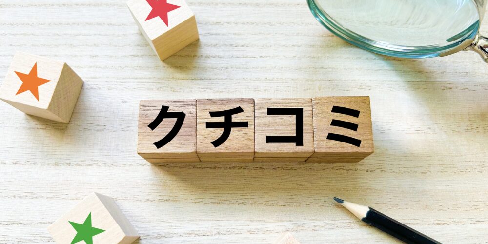 木目のテーブルに『クチコミ』と書かれた木製ブロックが並び、周囲に星マークのブロックや鉛筆、虫眼鏡が配置されている