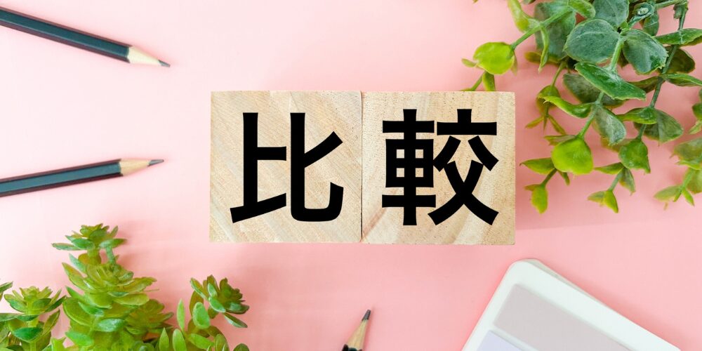 ピンクの背景に『比較』と書かれた木製ブロックを置き、周囲に観葉植物と鉛筆を配置したイメージ