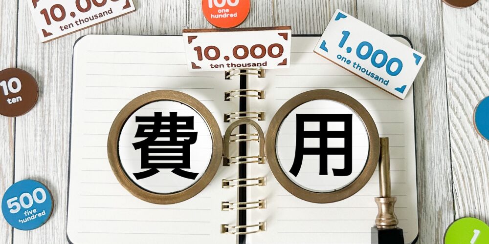 開いたリングノートの上に「費用」と「用」の文字を拡大鏡が強調し、周囲に「10000」「1000」「500」などの紙幣・硬貨アイコンが散りばめられた費用相場イメージ。