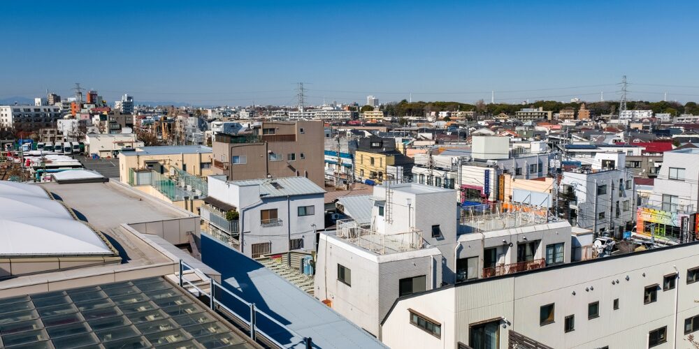 高い建物の屋上から見下ろした住宅街のパノラマ。密集する家屋と遠景のビル群、快晴の青空が広がっている。