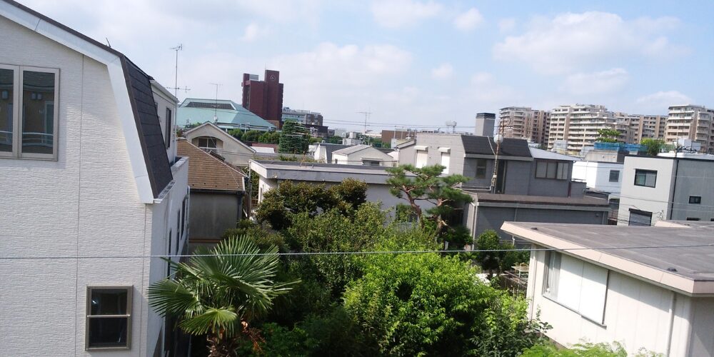 青空の下、緑豊かな庭木と住宅の屋根が並ぶ郊外の街並みを見渡す風景
