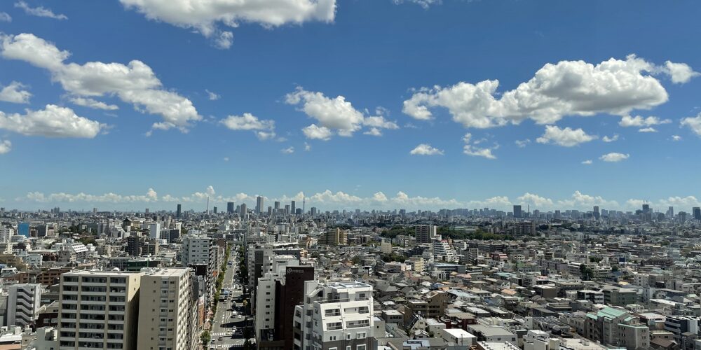 ビルやマンションが立ち並ぶ都市の街並みと、その上に広がる青空に白い雲が浮かんでいる景色。