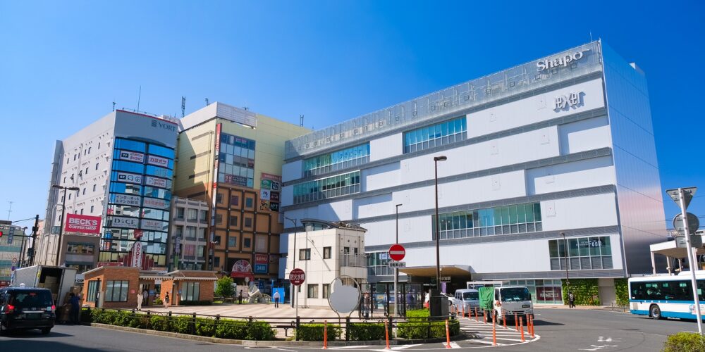 青空の下で大きなガラス張り商業施設「Shapo」と書かれた建物と、隣接する複数のビル・タクシー乗り場が並ぶ駅前風景。
