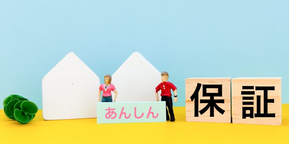 白い家型オブジェとミニチュアの男女、木製ブロック『保証』とプレート『あんしん』で安心保証を訴求するイメージ