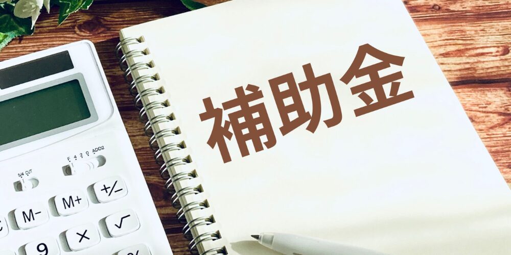 木目デスクに置かれた電卓とリングノート。ノートには大きく『補助金』の文字