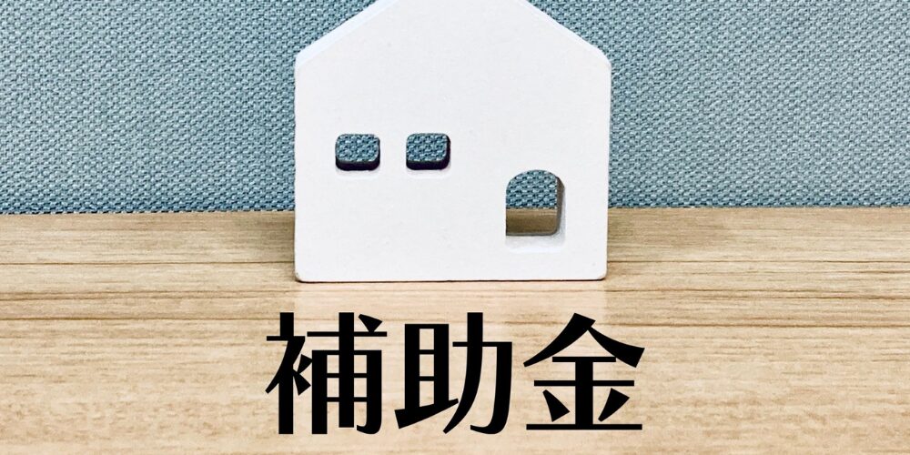 製テーブルに白い家型オブジェを置き、手前に『補助金』の文字を大きく配置したイメージ