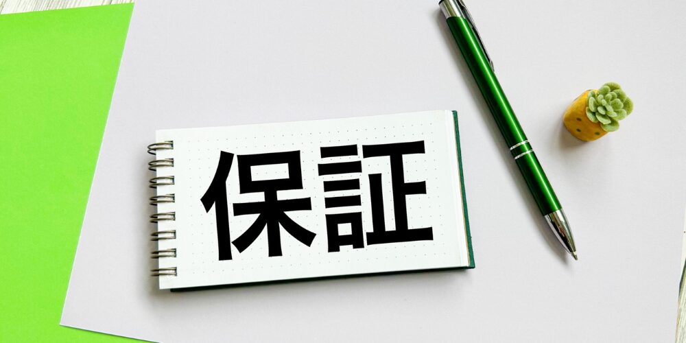 ノートパッドに「保証」と大きく書かれ、隣にペンと観葉植物が置かれたデスク上の様子