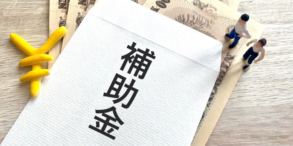 日本円の紙幣と黄色い円マークのオブジェが添えられた白い封筒に「補助金」と黒文字で書かれている。