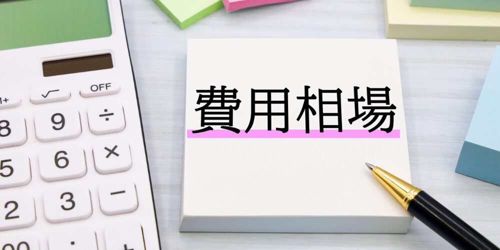 電卓と付箋の上に、下線付きで「費用相場」と黒字で書かれたメモが置かれたデスクまわりのクローズアップ。