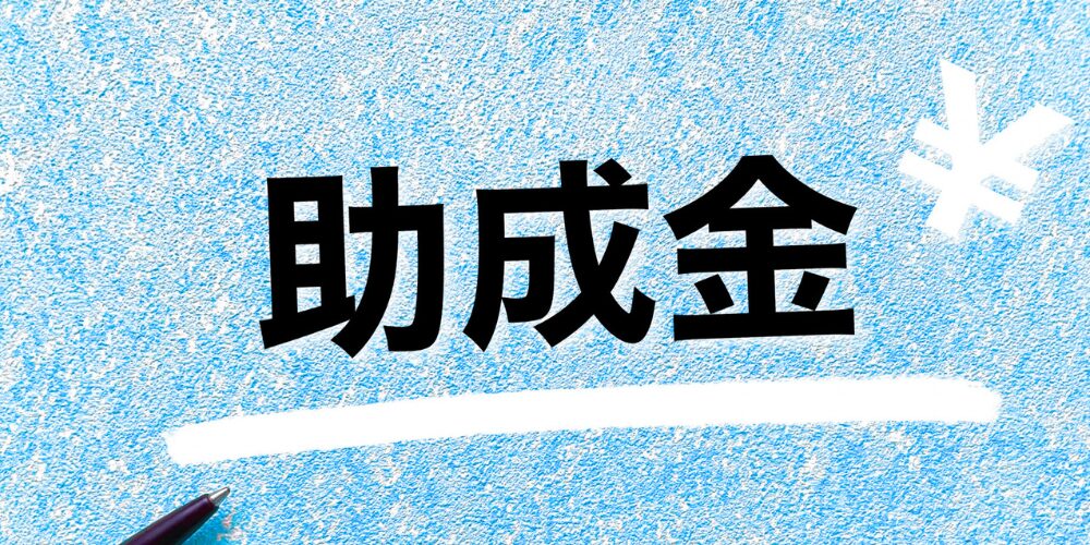 青いざらついた壁面に黒字で「助成金」と大きく書かれ、右端に白い円記号と下にペン先。