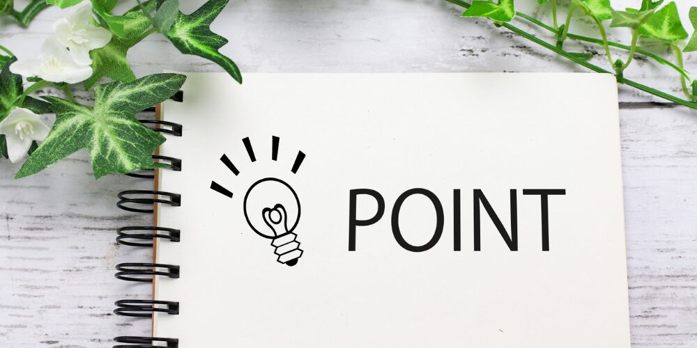 白いスパイラル綴じノートに電球のアイコンと「POINT」の文字が印刷され、周囲にグリーンの葉と文房具が配置されている。