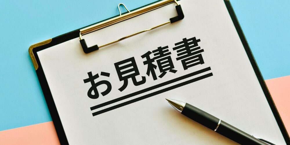 水色とピンクの背景にクリップボードを置き、黒いペン先のそばに太字で下線入りの「お見積書」と書かれた紙。