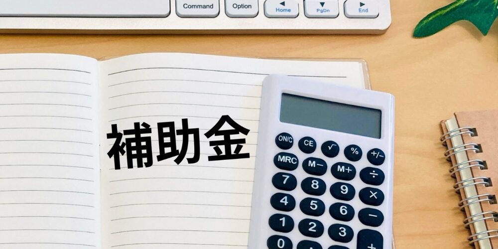 ノートパッドの見開きページに「補助金」と書かれ、その隣に電卓とキーボードの一部が写っている写真。