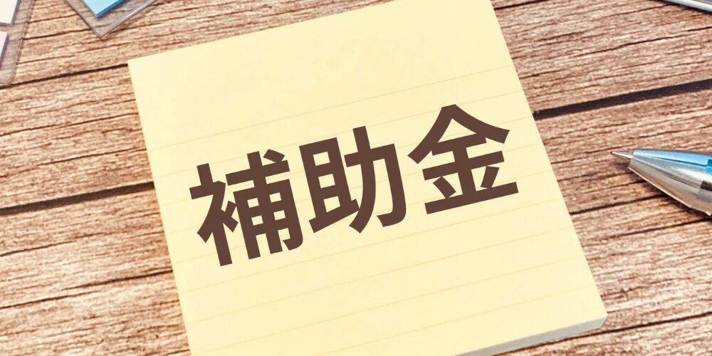 木製のデスク上に置かれた付箋に「補助金」と文字が書かれ、横にペンが添えられた写真。