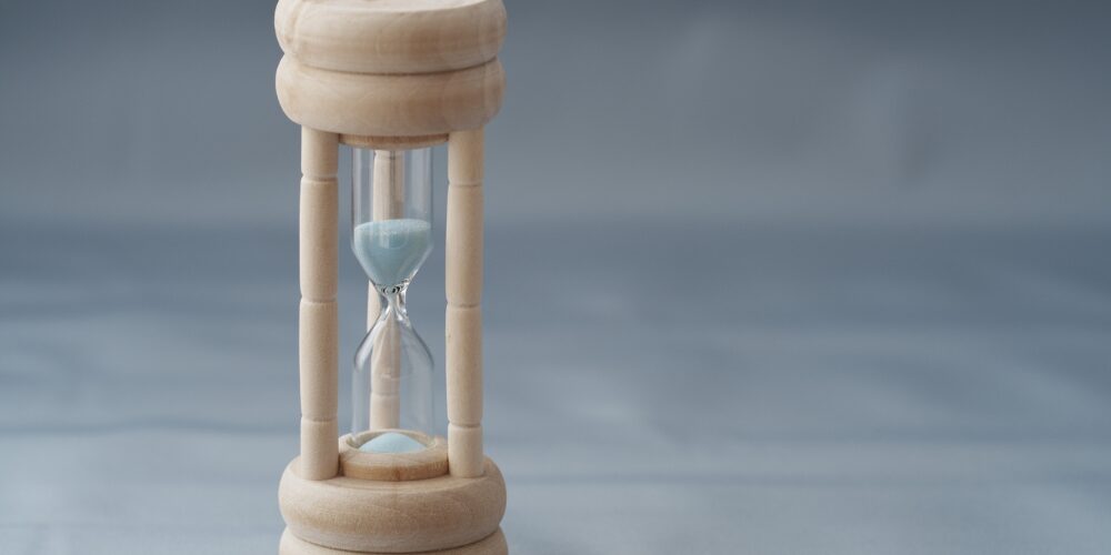 木製の枠に青い砂が入った砂時計。背景はぼかされ、時間の経過を表現している。