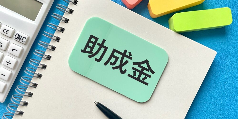 電卓と付箋の並ぶデスク上で、ノートに緑色のカードで「助成金」と書かれている様子